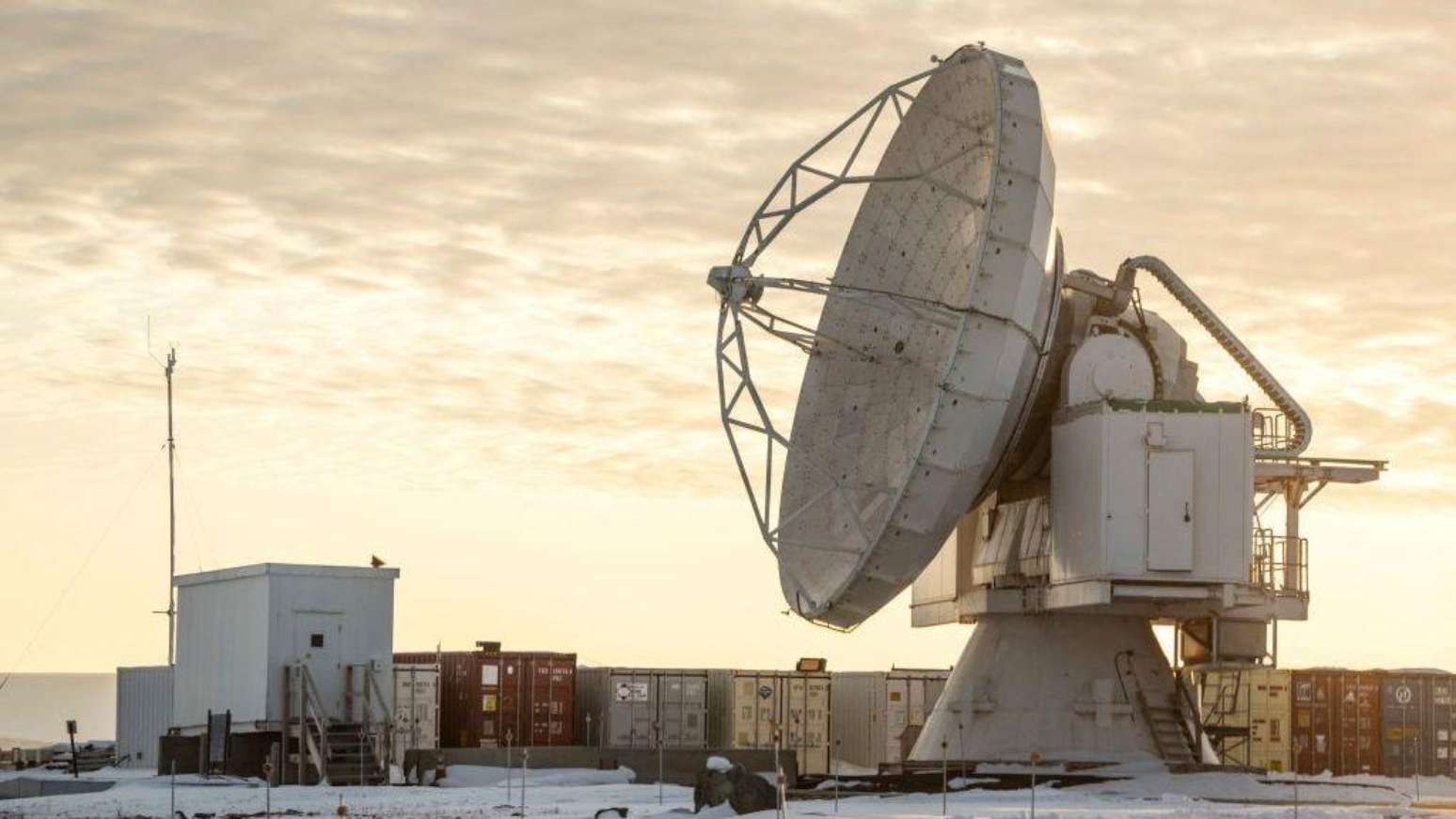 A Base Espacial Pituffik (antiga Base A&eacute;rea de Thule) &eacute; mostrada no norte da Groenl&acirc;ndia, em 4 de outubro de 2023. A imagem mostra uma grande antena parab&oacute;lica em frente a cont&ecirc;ineres, com um c&eacute;u alaranjado e nublado ao fundo.